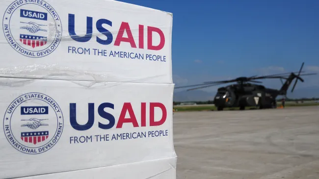 Boxes of aid from USAID are stacked on a tarmac near a US military helicopter